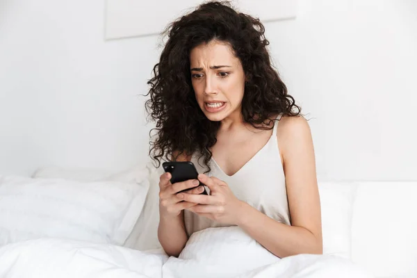 Portrait of displeased woman sitting in bed after sleep on white clean linen at bedroom with terrible emotions while using smartphone