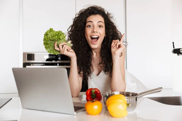 照片的快乐黑发妇女20S 持有绿色萨拉特和阅读食谱的笔记本电脑 而烹饪晚餐在厨房内饰 — 图库照片