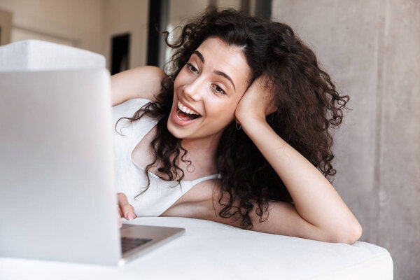 Photo closeup of young adorable woman with long curly hair wearing silk leisure clothing lying on couch in apartment and laughing while looking at screen of laptop