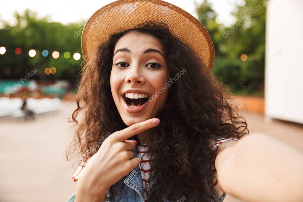Cheerful happy woman 18-20 with curly brown hair smiling and pointing finger aside while taking selfie photo outdoor