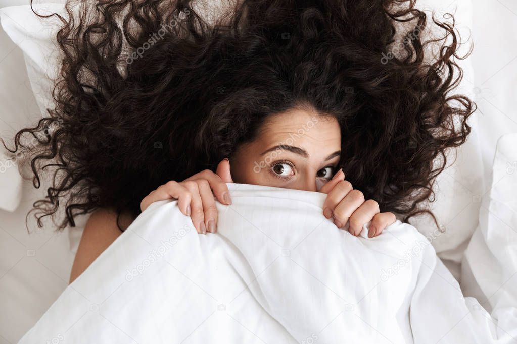 Portrait from above of cute woman 20s with dark curly hair covering her face with white blanket while lying in bed after waking up in morning