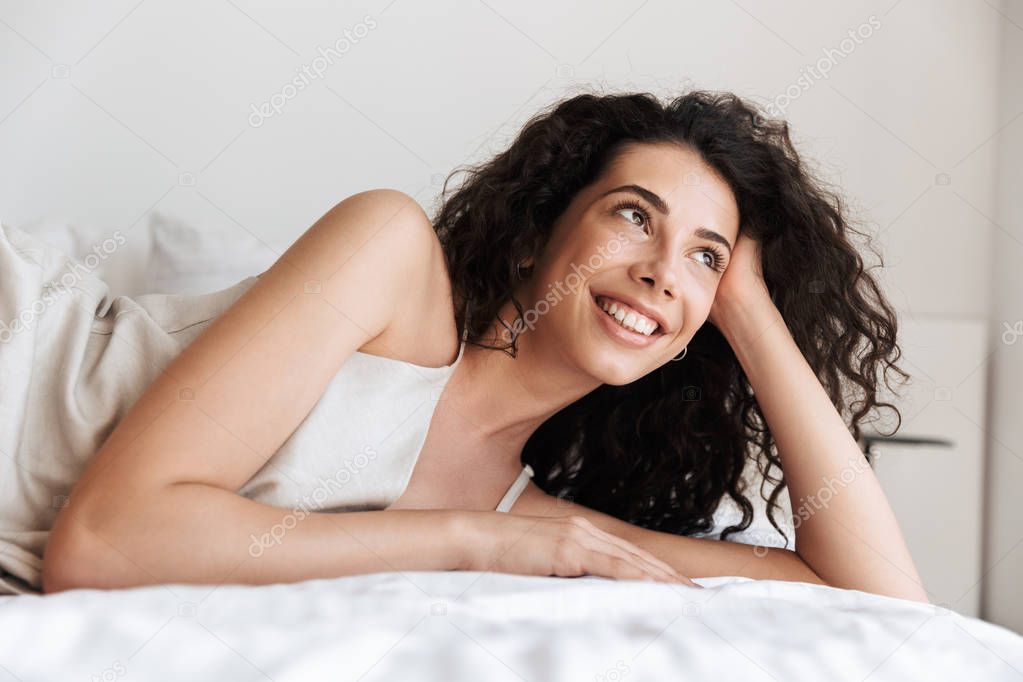 Closeup photo of pretty young woman 20s with long curly hair wearing silk leisure clothing lying in hotel bed and looking aside while propping up her head with hand
