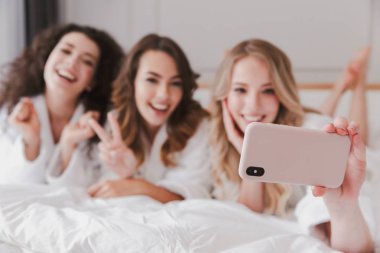Bulanık fotoğraf gülümseyen mutlu kadınların 20'li yaşlarda lüks yatak odalı evde ve alma selfie Smartphone cep telefonu ile tavuk parti sırasında yalan giyen beyaz bornoz