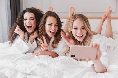 Üç Avrupa çok güzel kadınlar 20'li yaşlarda evde lüks yatak odasında yalan ve selfie fotoğraf üstünde hareket eden telefon sırasında gelin duş alarak beyaz bornoz giyiyor