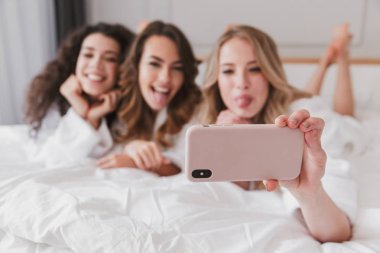 Neşeli kadınlar 20'li yaşlarda bulanık fotoğraf lüks daire ve alma selfie Smartphone cep telefonu ile rahat parlak yatak odasında tavuk parti sırasında yalan giyen beyaz bornoz