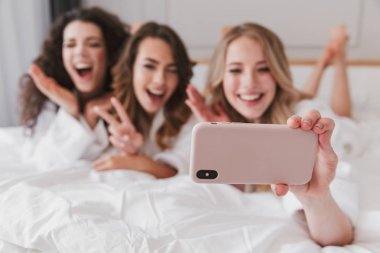 Bulanık fotoğraf gülümseyen mutlu kadınların 20'li yaşlarda lüks daire ve alma selfie Smartphone cep telefonu ile lüks yatak odasında tavuk parti sırasında yalan giyen beyaz bornoz