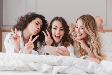 Mutlu fotoğraf gülümseyen güzel kadınların 20'li yaşlarda lüks yatak lüks daire içinde yalan ve selfie smartphone tavuk parti sırasında çekerken barış işareti gösteren beyaz bornoz giyiyor