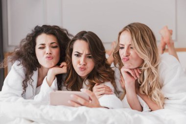 Üç genç kadın 20'li yaşlarda otel odasında şık yatak odasında yalan ve bekarlığa veda partisi sırasında cep telefonu selfie çekerken ekşitmeden beyaz bornoz giyiyor