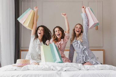 Sevindirici ve tavuk parti sırasında otel odasında rahat yatak üzerinde otururken elinde renkli alışveriş torbaları ile çığlık 20s giyen elbiseler güzel gülümseyen kadın mutlu fotoğraf