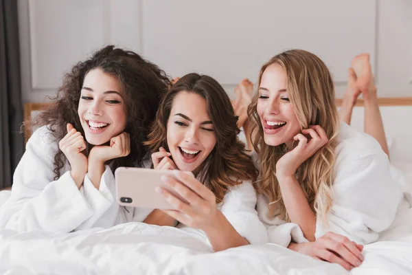 Happy photo of lovely smiling women 20s wearing white bathrobe lying in luxury bedroom in hotel room and taking selfie on cell phone during hen party