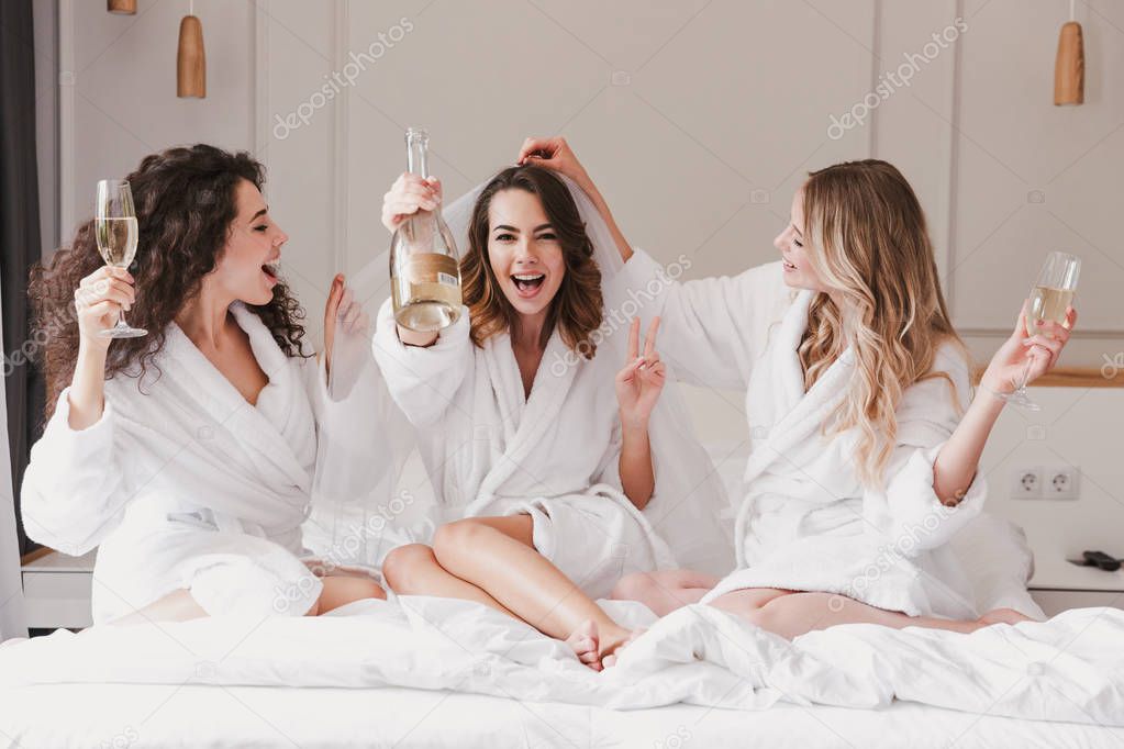 Portrait of pretty three women 20s celebrating bridal shower and drinking champagne while bride trying on wedding veil in posh apartment or hotel room