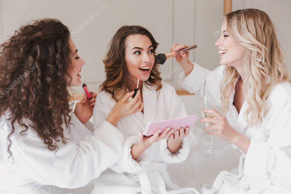 Young smiling women 20s wearing housecoat drinking champagne and applying makeup for bride during bridal shower while resting in luxury bedroom at hotel room