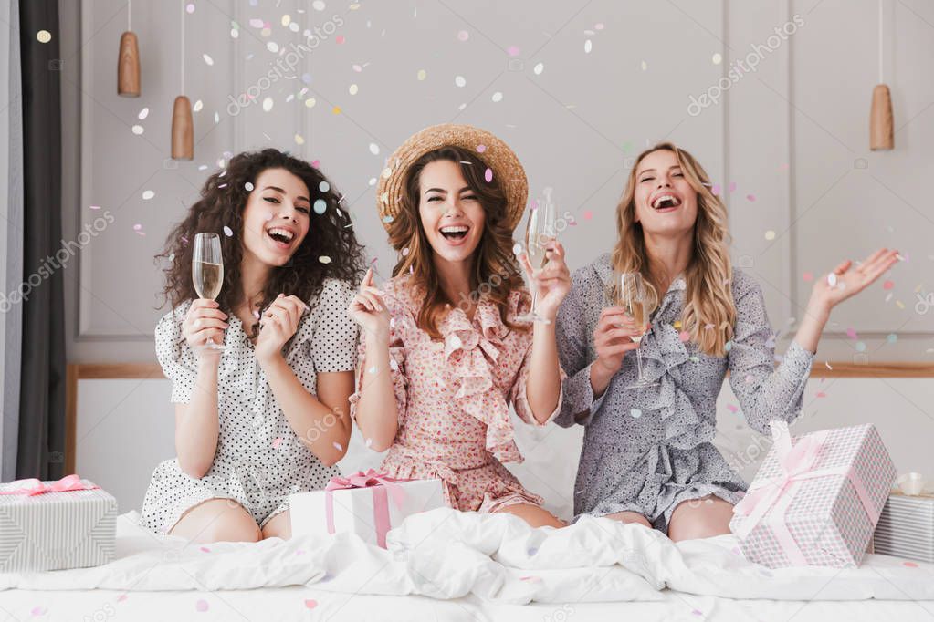 Portrait of beautiful excited women 20s wearing dresses celebrating bridal shower in posh apartment with champagne and falling confetti
