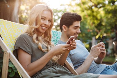 Mutlu güzel genç çift birlikte oturan şezlong üzerinde onların akıllı telefonlar açık havada kullanırken yan görünüm