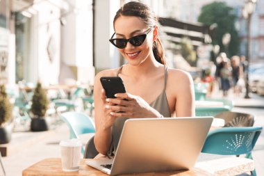 Yaz aylarında rahat kafe ya da restoran dışında otururken smartphone kullanarak siyah güneş gözlüğü takıyor mutlu kafkas kadın görüntüsünü