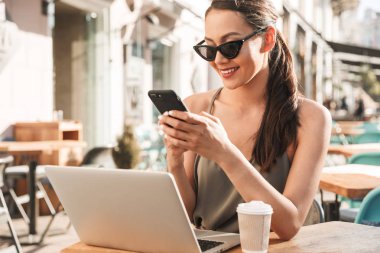 Görüntü güzel esmer kadının yaz aylarında rahat kafe ya da restoran dışında otururken elinde cep telefonu bakarak siyah güneş gözlüğü takıyor