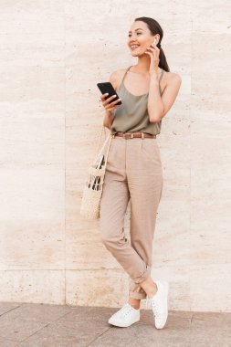 Bluetooth earpod ve holding cep telefonu elinde duran açık bej duvara dokunmak rahat yaz giyim Kafkas esmer kadın görüntüsünü