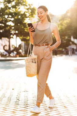 Şehir sokak yürüyüş ve hareket eden telefon tutarak trendy yaz kıyafeti giymiş esmer mutlu kadın fotoğrafı tam uzunlukta