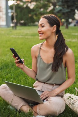 Dizüstü bilgisayar ve cep telefonu kullanarak açık havada Park çimenlerin üzerinde oturan pretty Asya kadın görüntüsünü.