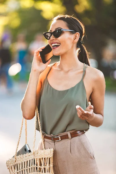 カジュアルな夏の服装と街を散策し 笑顔でスマート フォンで話す黒のサングラスを身に着けている美しいブルネットの女性のイメージ — ストック写真