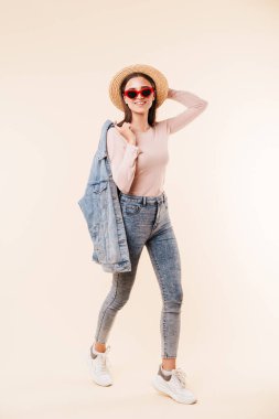 Tam uzunlukta bir süre ayakta ve beyaz arka plan üzerinde izole kamera bakarak poz hasır şapka ve denim ceket şık genç esmer kız portresi