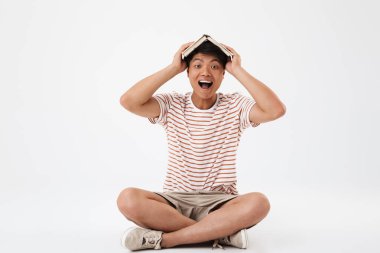 Beyaz arka plan üzerinde izole kafasına holding kitap çizgili tişört bacaklar ile katta otururken çığlık giyen memnun Asya adam portresi geçti