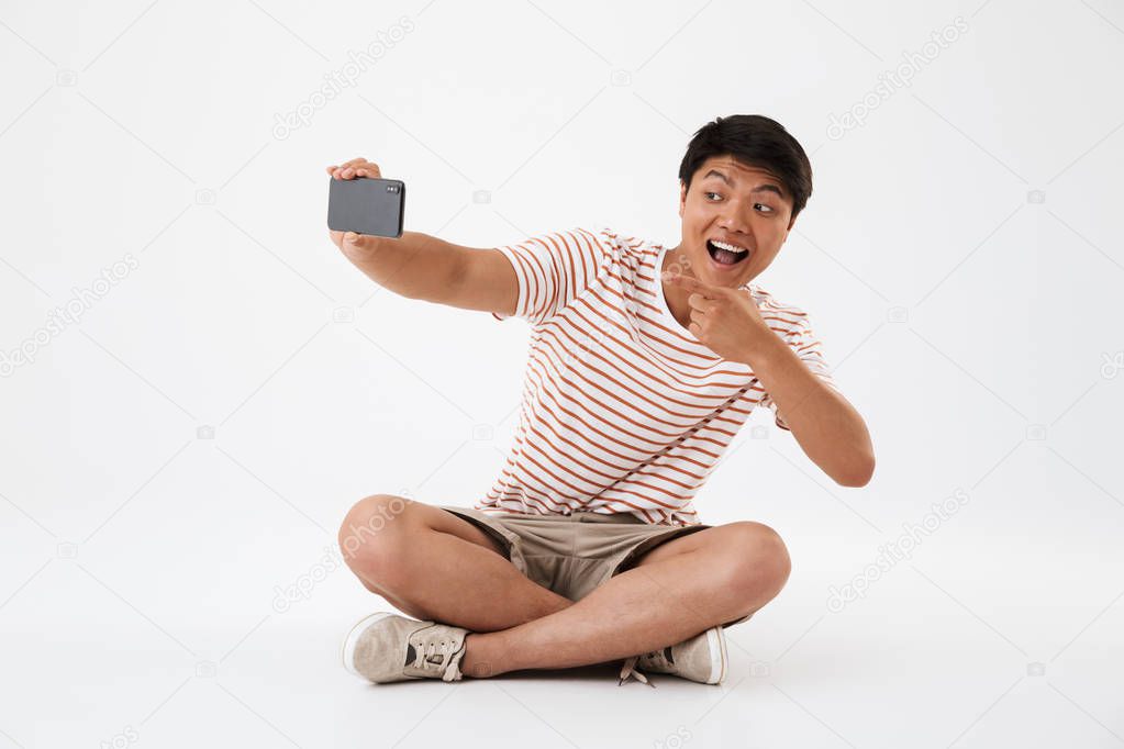 Portrait of a cheerful young asian man taking a selfie with mobile phone while sitting with legs crossed and showing thumbs up isolated over white
