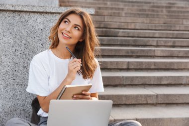 Genç kız holding defter ve kalem ile dizüstü bilgisayar açık havada merdivenlerde otururken gülümseyen
