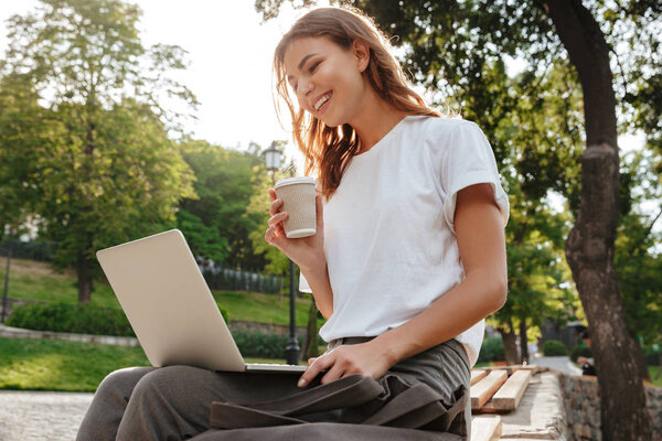 Image of caucasian modern woman sitting on bench in green park on summer day and using silver laptop while drinking takeaway coffee from paper cup