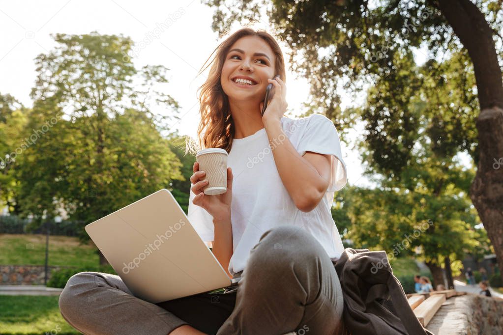 Image of smiling modern woman sitting on bench in green park on summer day and talking on smartphone while using silver laptop and drinking takeaway coffee