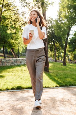 Tam boy görüntü yeşil parkın içinden dar yol aşağı yaz gününde yürüyüş ve paket servisi olan restoran kahve ile Smartphone cep telefonu ile konuşurken beyaz gülümseyen kadın