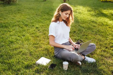 Fotoğraf laptop ve bluetooth earpod kullanırken yaz günü sırasında bacakları ile parkta yeşil çimenlerin üzerinde oturan Avrupa kadın 20'li yaşlarda geçti