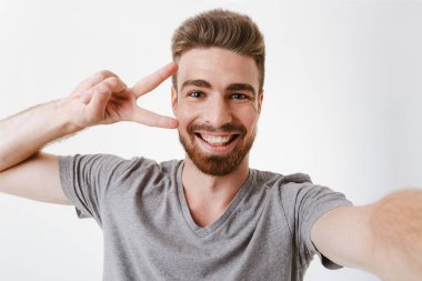 Beyaz arka plan üzerinde izole bir selfie çekerken barış hareketi gösterilen neşeli Genç sakallı bir adam portresi