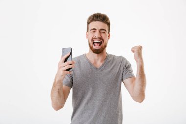 Kazanan jest yapmak imge-in heyecanlı genç adam cep telefonuyla beyaz duvar arka plan üzerinde izole ayakta.