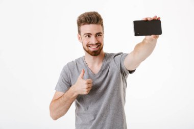 Hareket eden telefon ile jest Yaşasın tarafından selfie yapmak imge-in mutlu yakışıklı genç adam beyaz duvar arka plan üzerinde izole ayakta.