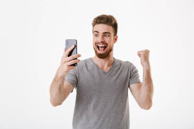 Kazanan jest yapmak imge-in heyecanlı genç adam cep telefonuyla beyaz duvar arka plan üzerinde izole ayakta.