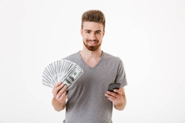 Genç adam cep telefonuyla para tutan beyaz duvar arka plan üzerinde izole ayakta görüntüsü. Görünümlü kamera.