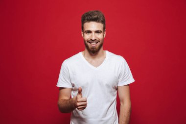 Neşeli yakışıklı genç başparmak kırmızı arka plan üzerinde izole göstermek yukarıya beyaz t-shirt görünümlü kamera görüntüsünü.