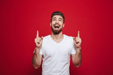 Görüntü izole kırmızı arka plan işaret üzerinde beyaz t-shirt yakışıklı mutlu genç adam. Bir kenara arıyorsunuz.