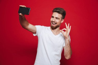 Neşeli bir genç sakallı adam ok hareket izole kırmızı arka plan üzerinde bir selfie çekerken gösterilen portresi
