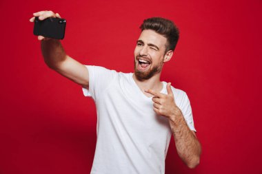 Kırmızı arka plan üzerinde izole bir selfie fotoğraf çekerken neşeli Genç sakallı adam işaret eden bir parmağa portresi