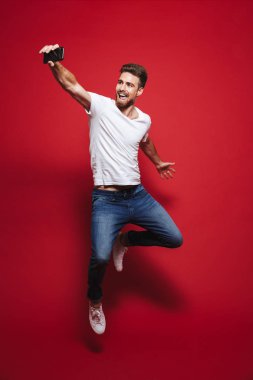 Neşeli bir genç sakallı adam başarı kutluyor ve atlama izole kırmızı arka plan üzerinde iken bir selfie alarak tam uzunlukta portresi