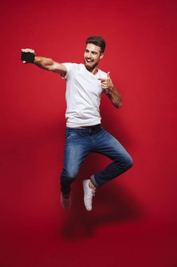 Neşeli bir genç sakallı adam başarı kutluyor ve atlama izole kırmızı arka plan üzerinde iken bir selfie alarak tam uzunlukta portresi
