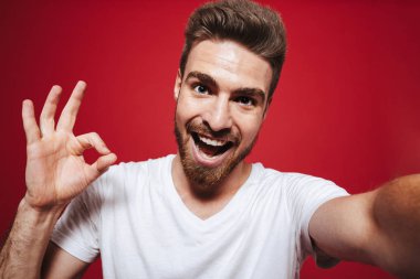 Neşeli bir genç sakallı adam ok hareket izole kırmızı arka plan üzerinde bir selfie çekerken gösterilen portresi