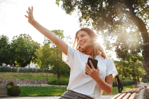 Image of smiling friendly woman sitting on bench in green park on summer day and waving hand aside saying hello while holding mobile phone