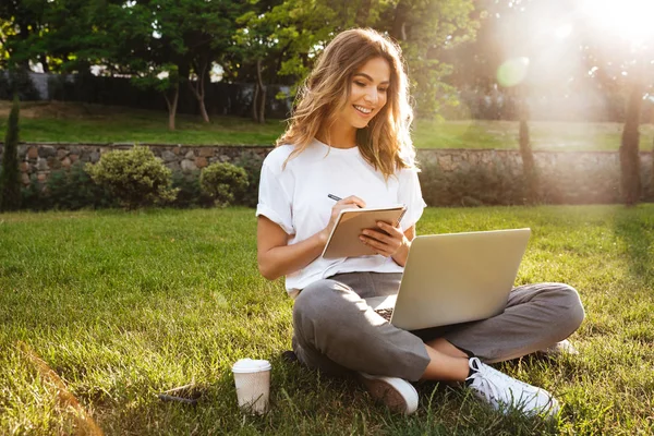 有魅力的年轻女子的肖像坐在公园的绿色草地上 腿横渡在夏天的时间和写笔记与钢笔 而使用笔记本电脑 — 图库照片