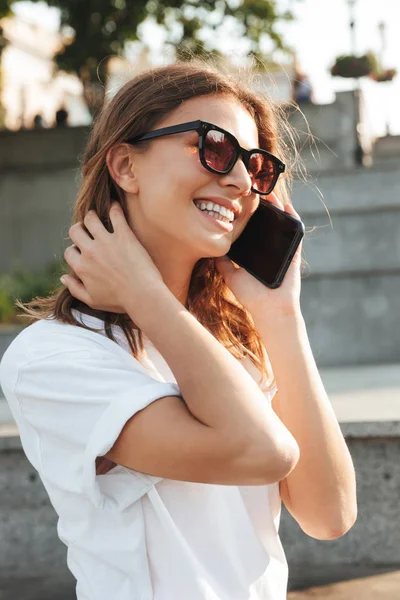 欧洲快乐女子穿着休闲夏季套装和太阳镜笑着走在城市街道上 微笑着谈论智能手机的照片 — 图库照片