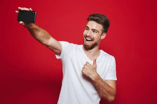 赤い背景に分離された Selfie しながらジェスチャー親指を示す陽気な若いひげを生やした男の肖像 — ストック写真