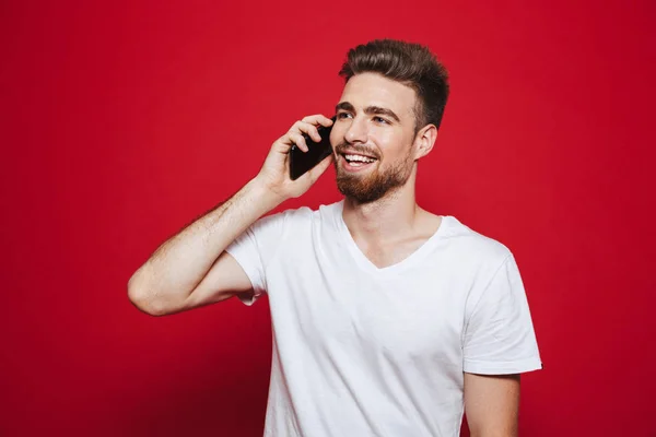赤い背景に分離された携帯電話で話している幸せな若いひげを生やした男の肖像 — ストック写真