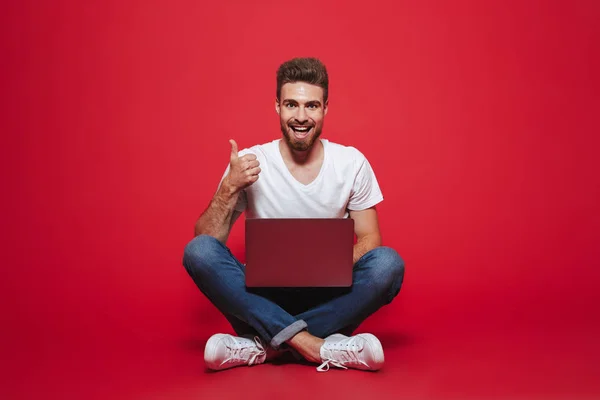一个快乐的年轻胡子男子的肖像显示竖起大拇指的手势 而坐在笔记本电脑孤立的红色背景 — 图库照片